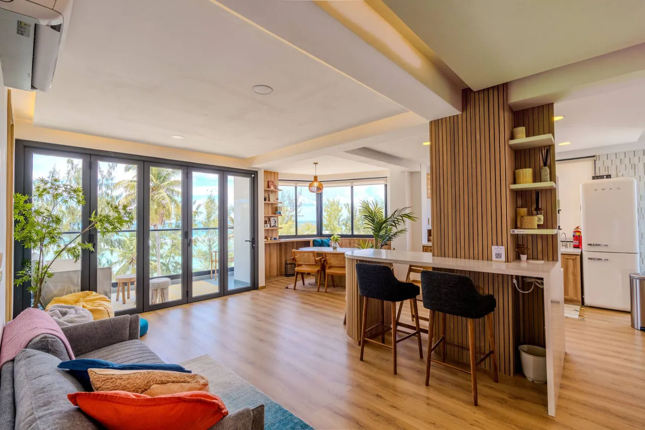 Open-plan living room with a sofa, a timber bar counter with stools and floor-to-ceiling windows onto palms and the sea