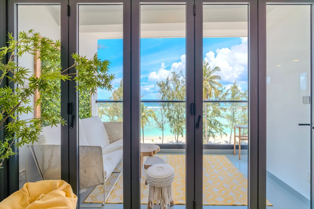 Glass doors open onto the balcony, with palms and the turquoise sea beyond