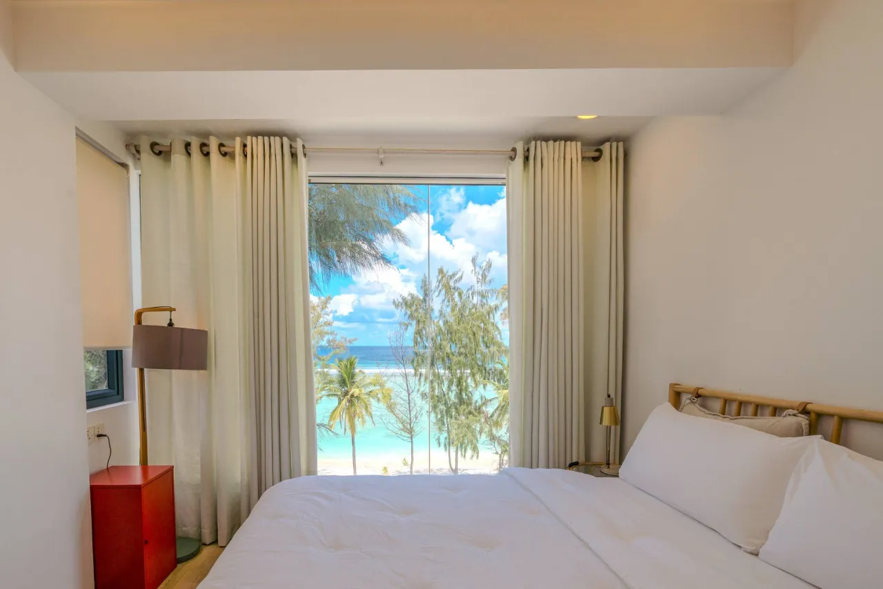 Large white bed facing a curtained window, with palms and turquoise sea outside